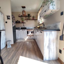 Stolarstwo Janicki Henryk - Kuchnia w stylu rustykalnym z białymi, postarzanymi frontami szafek i czarnym blatem. Widoczne drewniane półki z dekoracjami i roślinami. Ciepłe światło podkreśla naturalny charakter wnętrza.