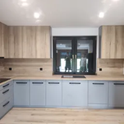 Nowoczesna kuchnia na wymiar z szarymi szafkami i drewnopodobnym blatem, czarne uchwyty, okno z ciemną ramą, drewniana podłoga. W oknie odbija się osoba.