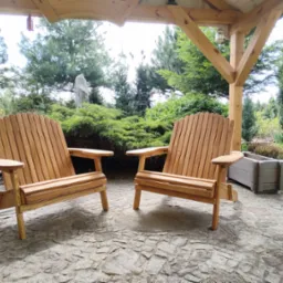 Dwa drewniane krzesła Adirondack na kamiennej posadzce pod drewnianą altaną, z piecem kaflowym po lewej i zielenią w tle. Jasne drewno, naturalne światło.