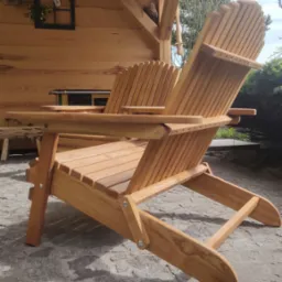 Drewniany fotel ogrodowy typu Adirondack, wykonany z jasnego drewna, ustawiony na kamiennej posadzce, z zadaszeniem w tle. Widoczny detal wykonania i naturalna faktura drewna.