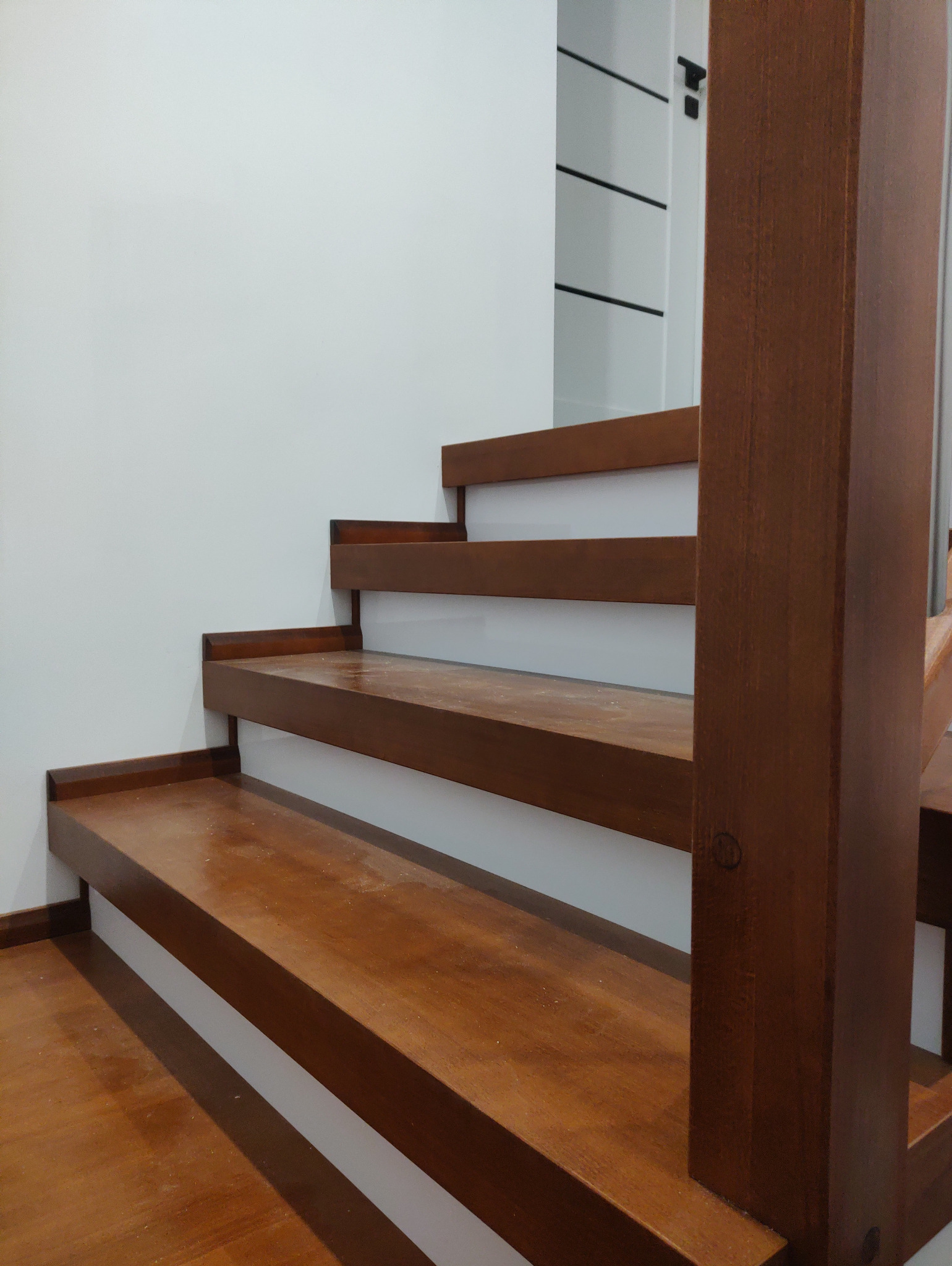 Nowoczesne schody drewniane z ciemnego drewna, z białymi podstopnicami, widoczne z dołu, z elementami balustrady. Minimalistyczny design i oświetlenie.