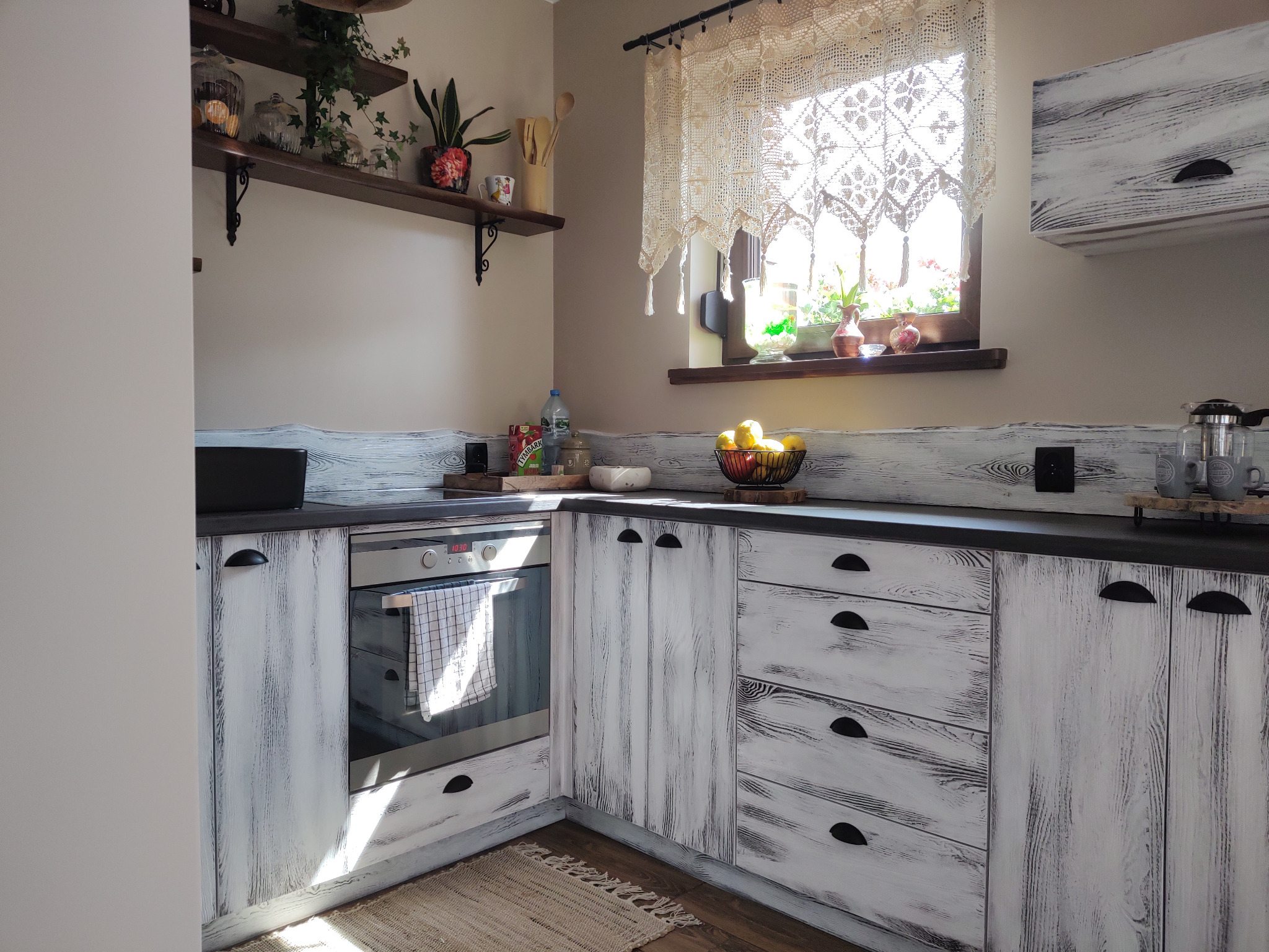 Rustykalna kuchnia z białymi, postarzanymi frontami. Blat w kolorze ciemnego grafitu. Na blacie owoce, przyprawy i dekoracje. Okno zdobi koronkowa firanka.