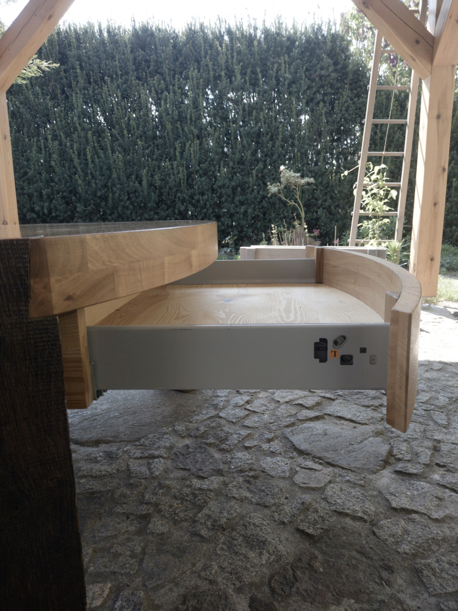 Otwarta szuflada z jasnego drewna w konstrukcji altany, z widocznym mechanizmem. Kamienna podłoga i drabina w tle.