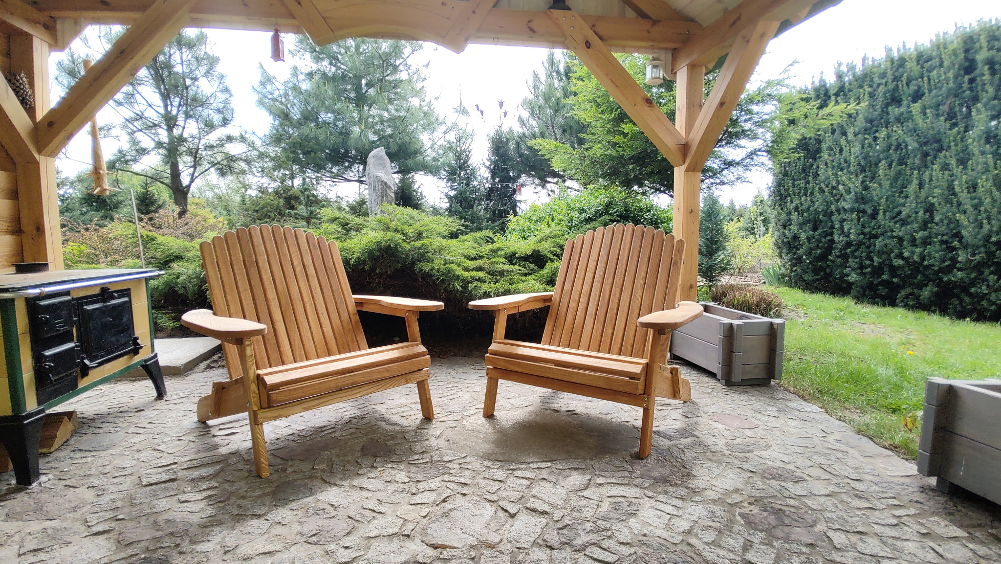 Dwa drewniane krzesła Adirondack na kamiennej posadzce pod drewnianą altaną, z piecem kaflowym po lewej i zielenią w tle. Jasne drewno, naturalne światło.