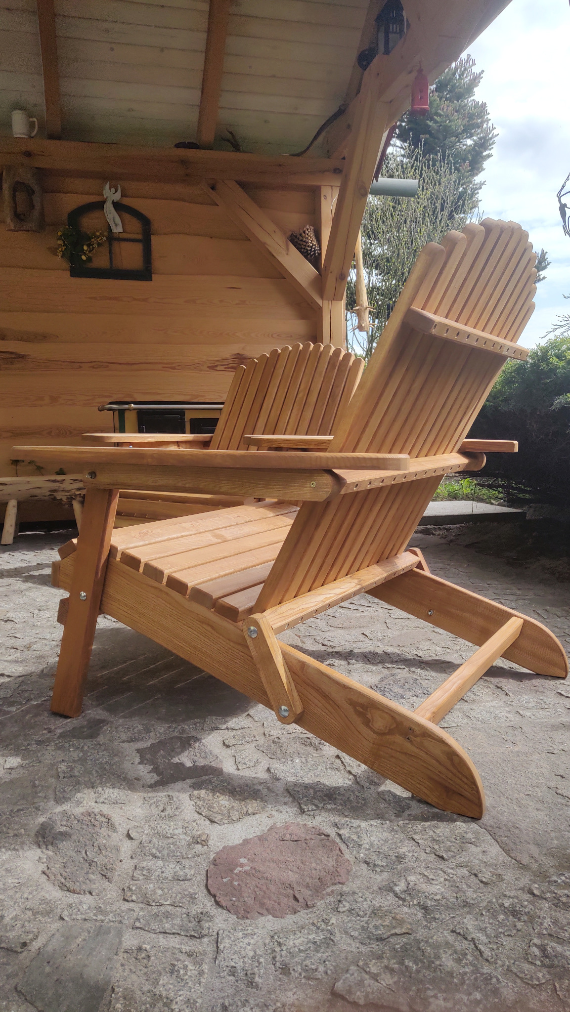 Drewniany fotel ogrodowy typu Adirondack, wykonany z jasnego drewna, ustawiony na kamiennej posadzce, z zadaszeniem w tle. Widoczny detal wykonania i naturalna faktura drewna.