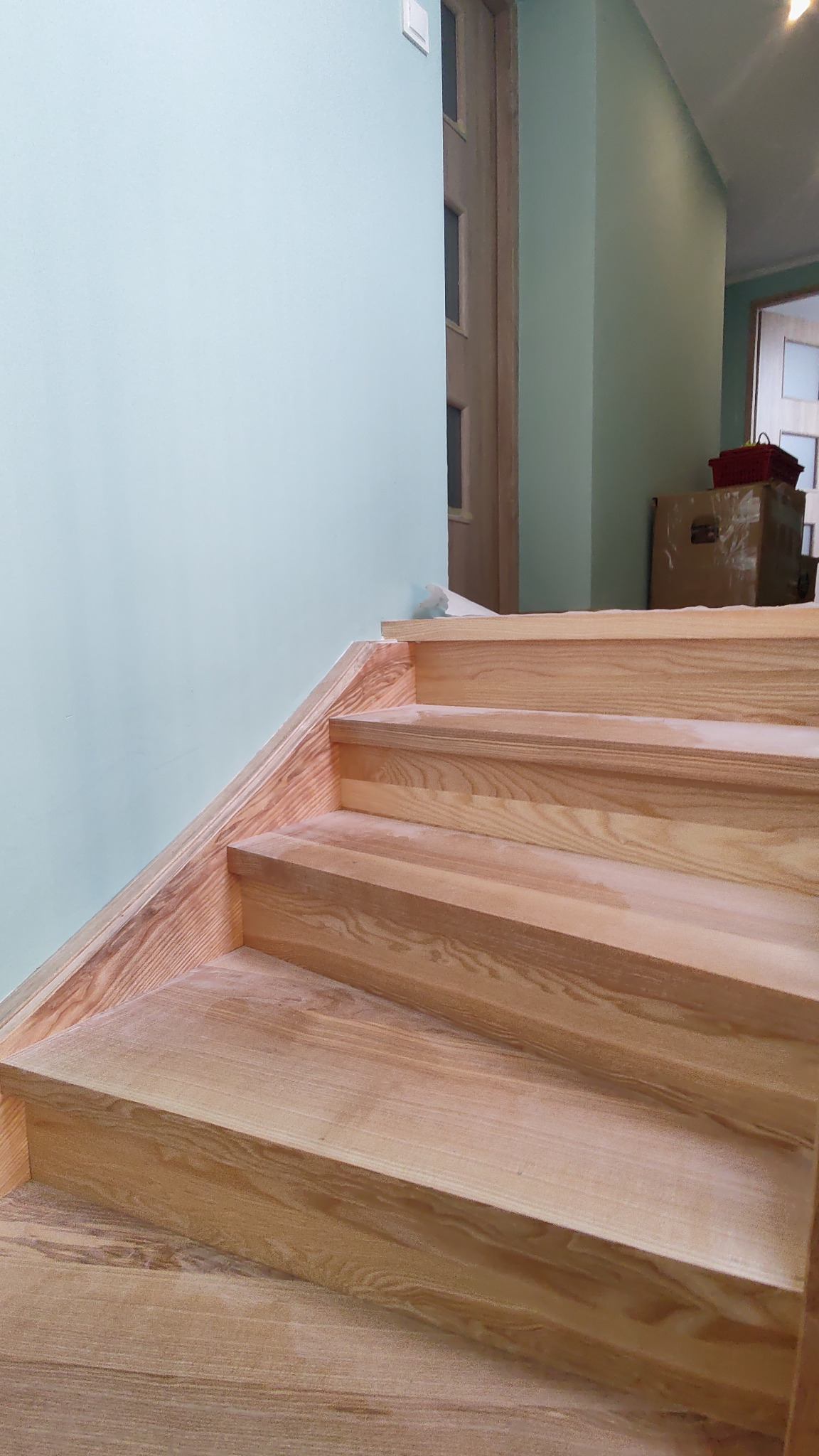 Jasne, drewniane schody z widocznym usłojeniem, prowadzące w górę, z błękitną ścianą w tle. Widoczne detale wykończenia i naturalna kolorystyka drewna.
