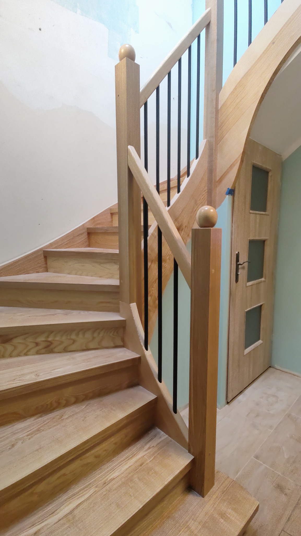 Jasne, drewniane schody z czarnymi balustradami i ozdobnymi gałkami, prowadzące w górę. Widoczny fragment drzwi z szybkami i jasna podłoga.