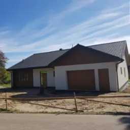 Nowoczesny dom parterowy z garażem w trakcie wykończenia, widok z zewnątrz, błękitne niebo z delikatnymi chmurami, teren przygotowany pod trawnik.