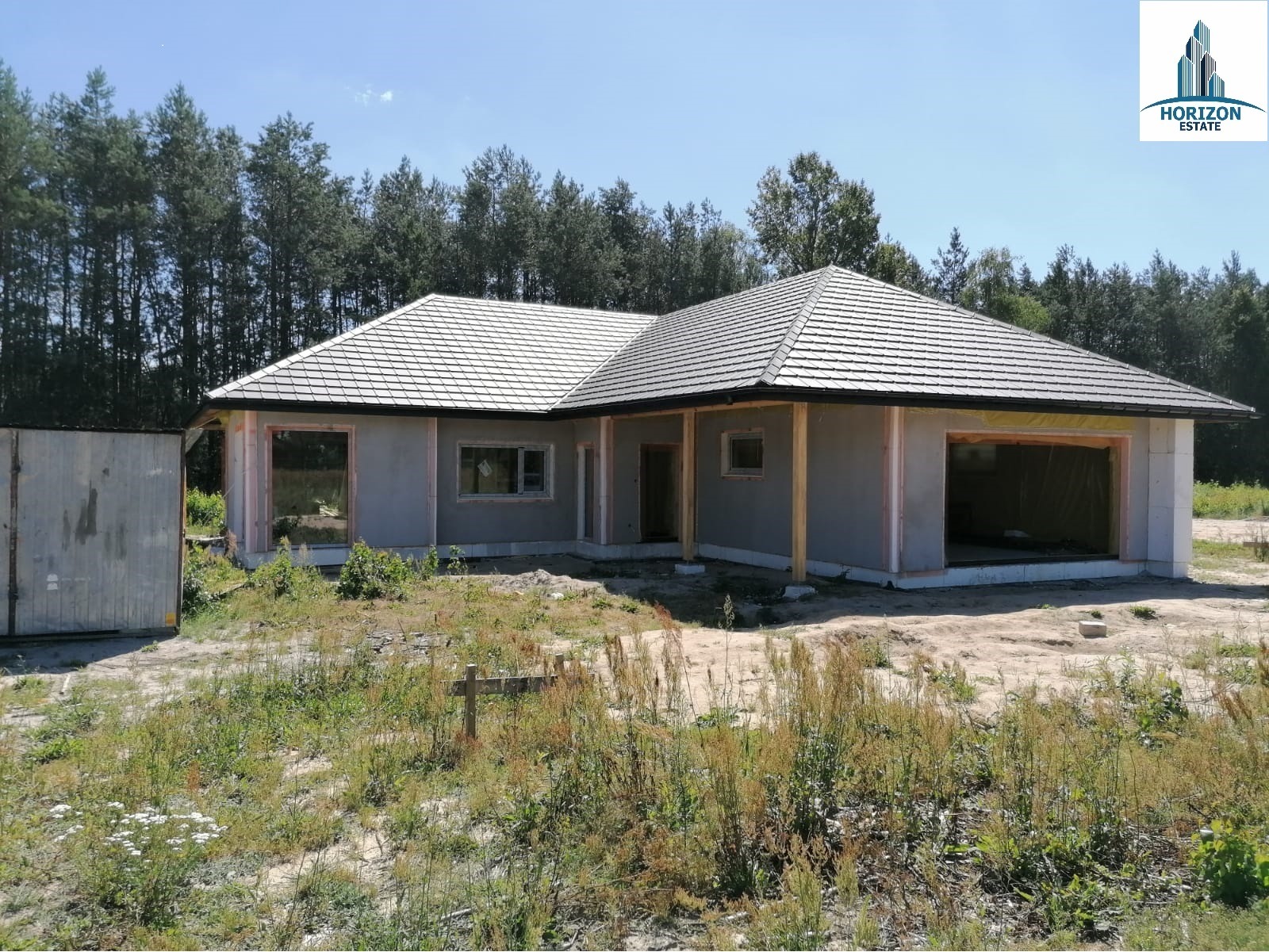 Niedokończony parterowy dom z szarą elewacją i ciemną dachówką na tle lasu. Widoczne niezamontowane okna i drzwi. Na pierwszym planie zarośnięty teren.