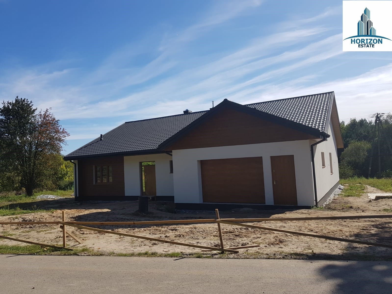 Nowoczesny dom parterowy z garażem w trakcie wykończenia, widok z zewnątrz, błękitne niebo z delikatnymi chmurami, teren przygotowany pod trawnik.