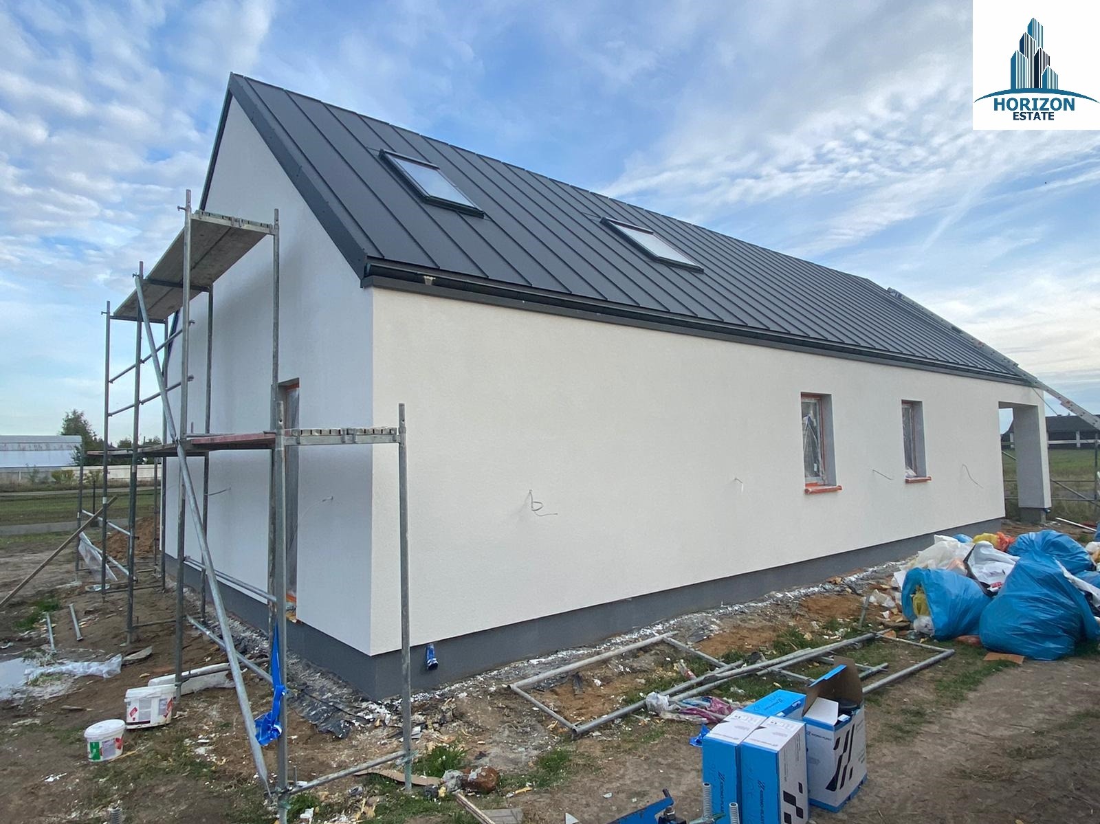 Boczny widok domu w trakcie budowy: białe ściany, grafitowy dach z oknami połaciowymi, rusztowania i materiały budowlane wokół. Widoczne logo Horizon Estate.