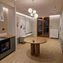 Rubi-Bud - Jasne wnętrze z parkietem w jodełkę, okrągłym stołem i elementami dekoracyjnymi. Widoczna kuchnia z zabudową w odcieniach zieleni i przeszklone drzwi balkonowe.