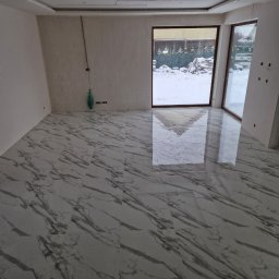 Rubi-Bud - Wykończona podłoga z płytek imitujących marmur w jasnym pomieszczeniu z dużymi oknami. Widoczne niedokończone ściany i sufit podwieszany.