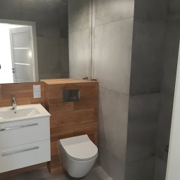 Rubi-Bud - Nowoczesna łazienka z szarymi płytkami, drewnianą zabudową WC i białą szafką z umywalką. Lustro z oświetleniem LED odbija fragment wnętrza.