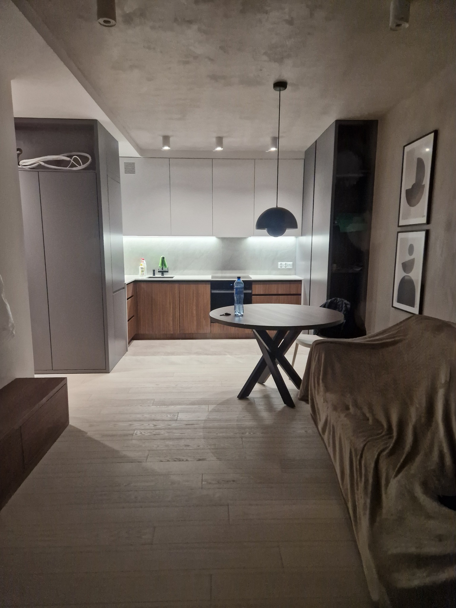 Nowoczesne wnętrze mieszkania: drewniana podłoga, zabudowa kuchenna z drewnianymi frontami, okrągły stół z butelką wody, minimalistyczne obrazy na ścianie.