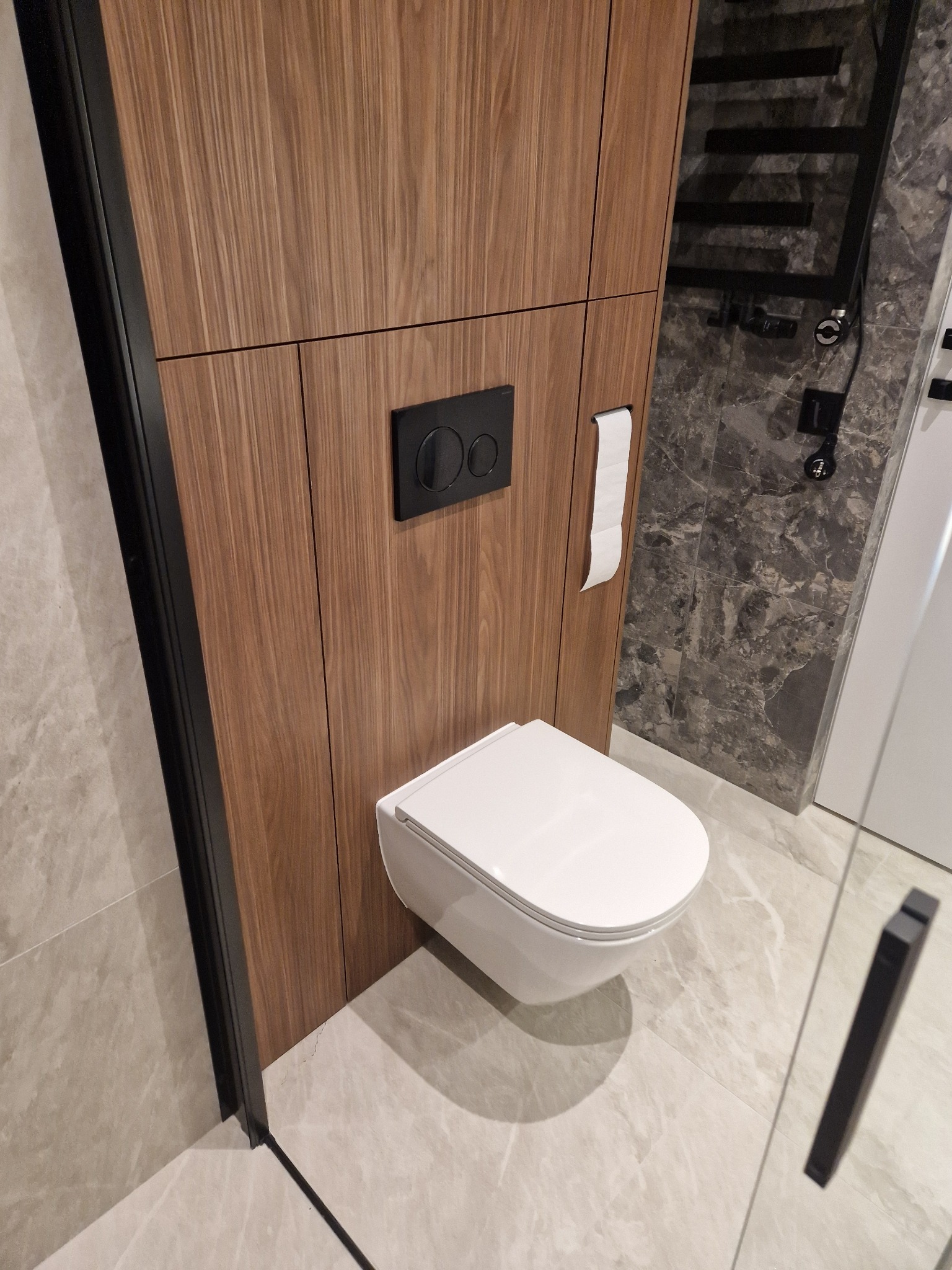 Nowoczesna toaleta podwieszana z drewnianą zabudową i czarnym przyciskiem spłukującym, marmurowe płytki na ścianie i podłodze. Minimalistyczny design łazienki.