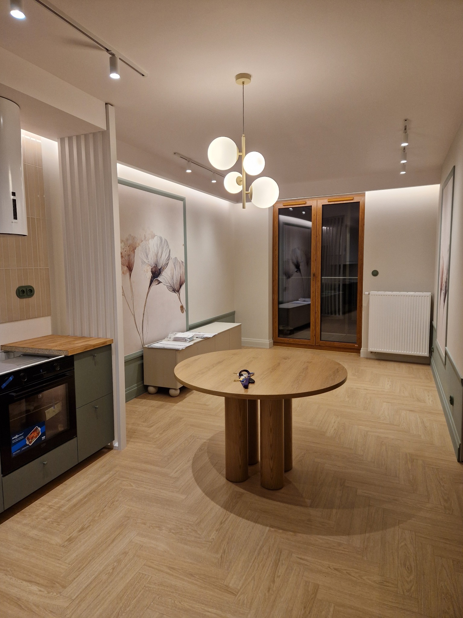 Jasne wnętrze z parkietem w jodełkę, okrągłym stołem i elementami dekoracyjnymi. Widoczna kuchnia z zabudową w odcieniach zieleni i przeszklone drzwi balkonowe.
