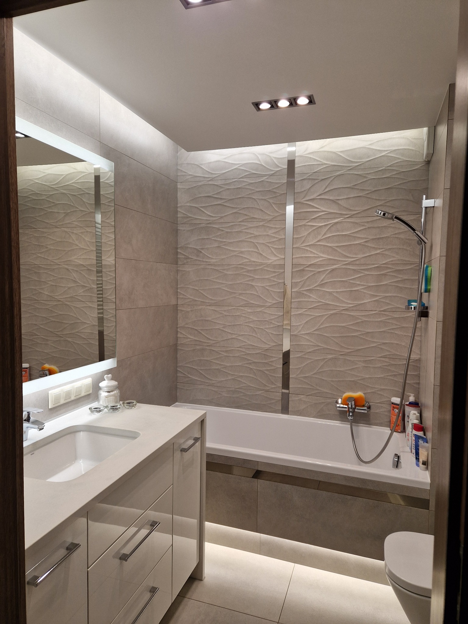 Nowoczesna łazienka z podświetleniem LED pod szafką umywalkową i wanną, beżowe płytki z falistym wzorem na ścianach, lustro z oświetleniem i biała armatura.