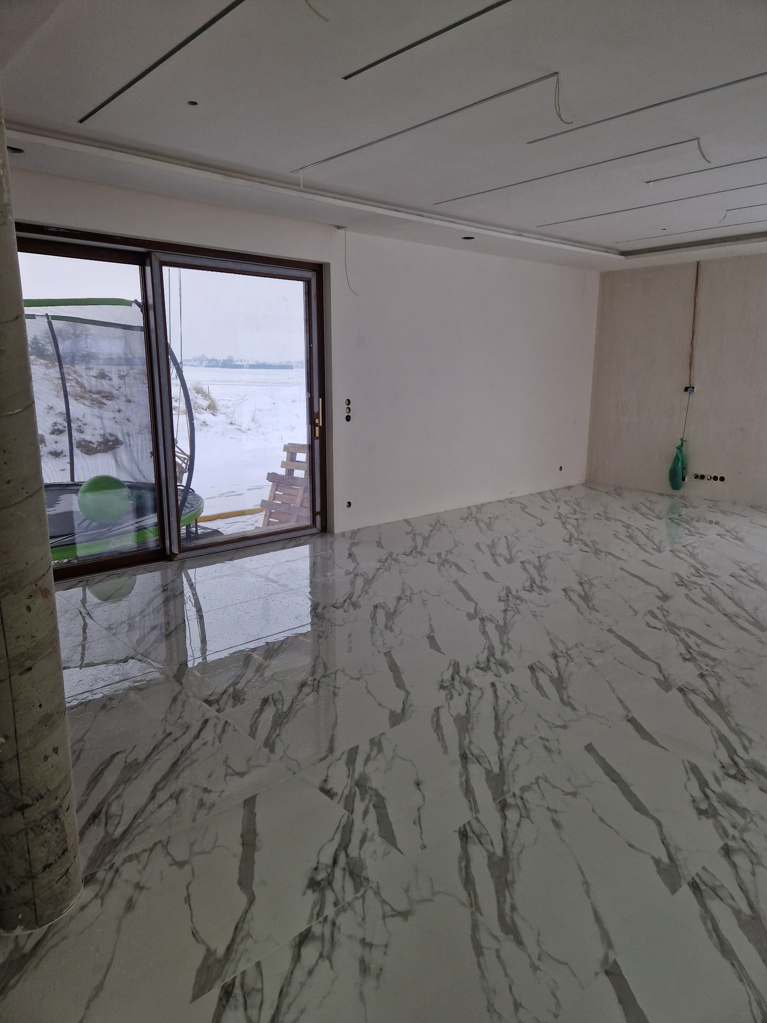 Jasne, przestronne wnętrze z marmurową podłogą i dużym oknem balkonowym. Widoczne niedokończone prace na suficie i ścianach. Za oknem zimowy krajobraz.