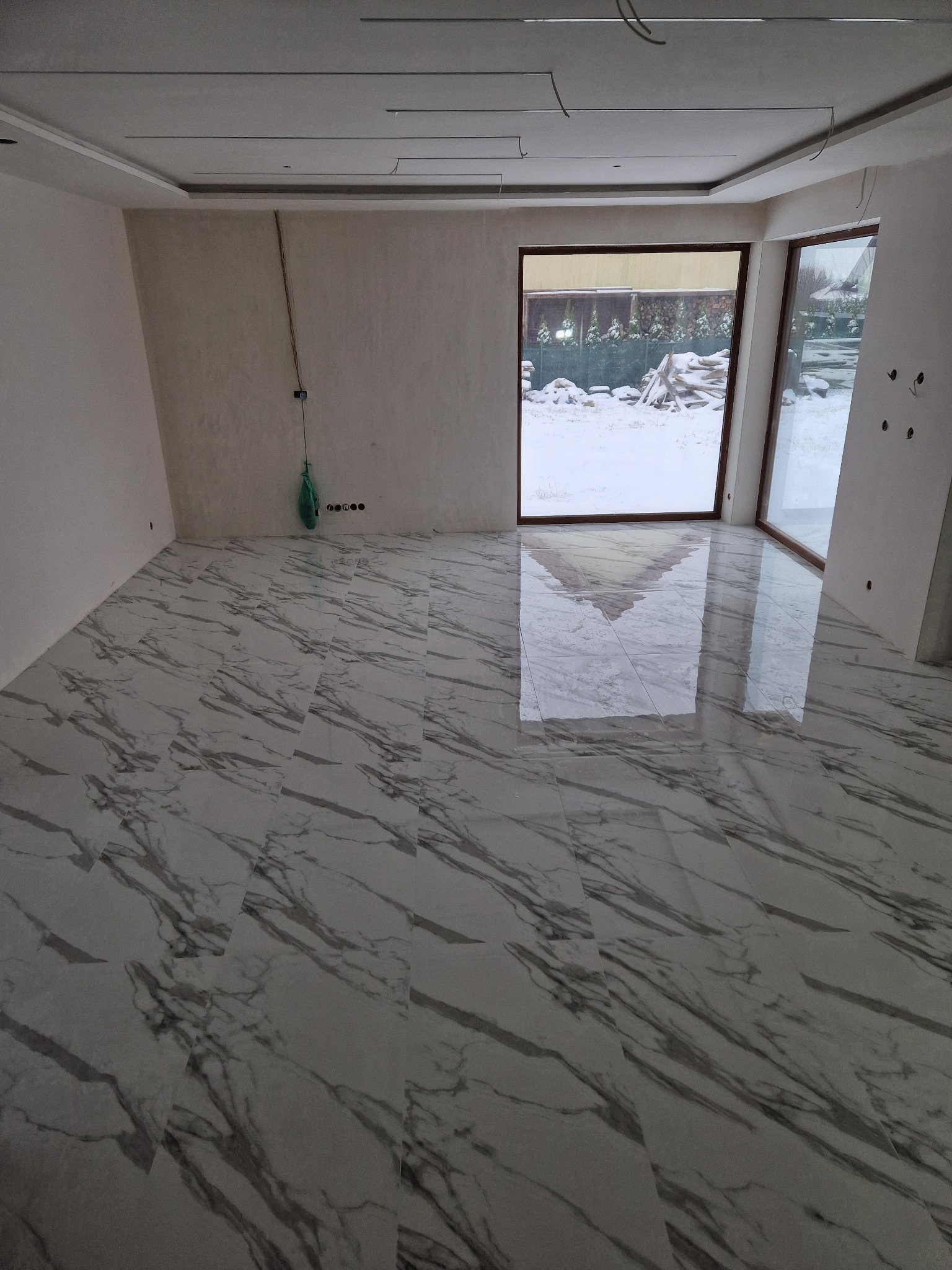 Wykończona podłoga z płytek imitujących marmur w jasnym pomieszczeniu z dużymi oknami. Widoczne niedokończone ściany i sufit podwieszany.