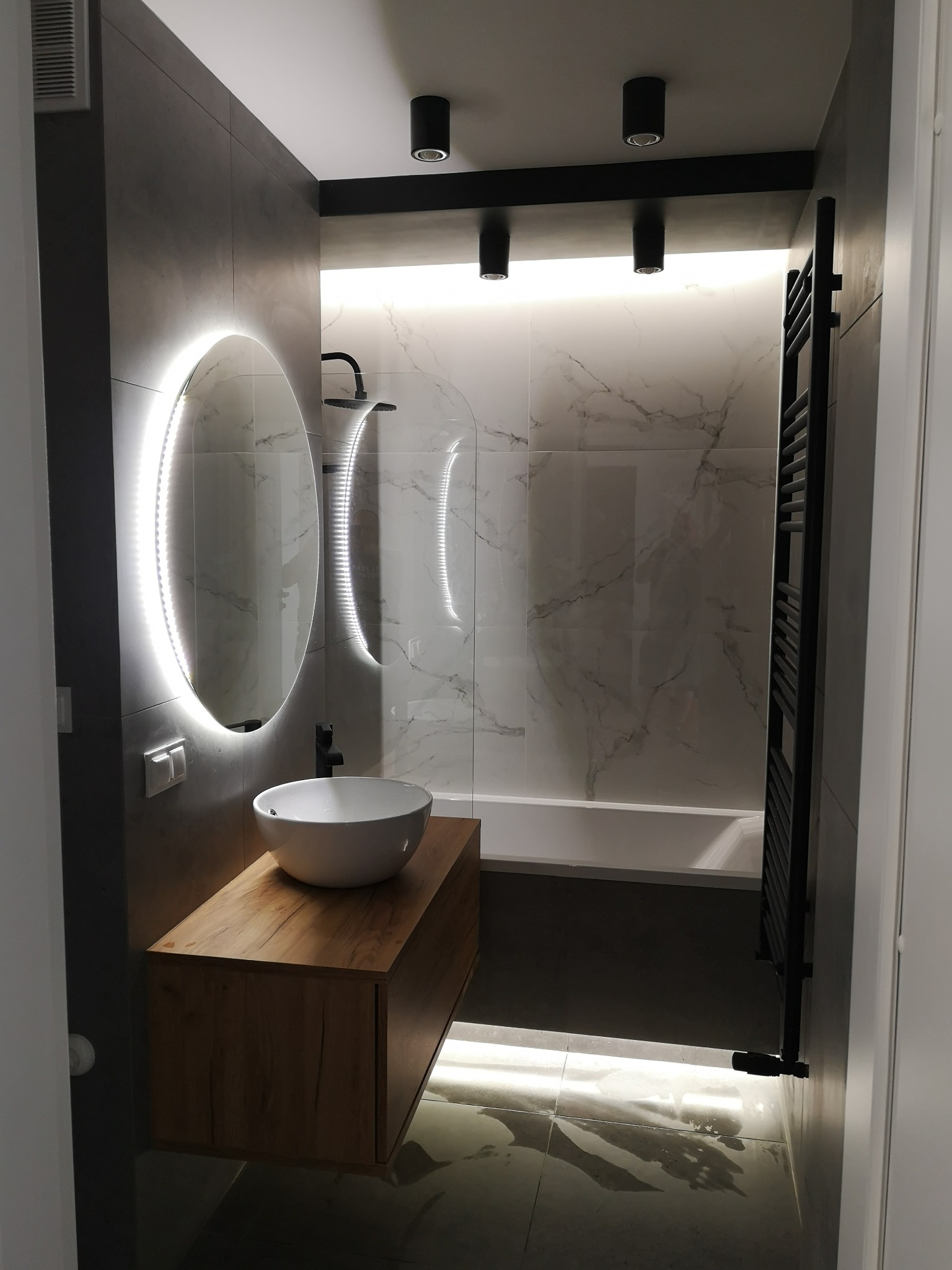 Nowoczesna łazienka z owalnym lustrem LED, drewnianą szafką pod umywalką i marmurowymi płytkami. Czarne akcenty oświetlenia i grzejnika drabinkowego.