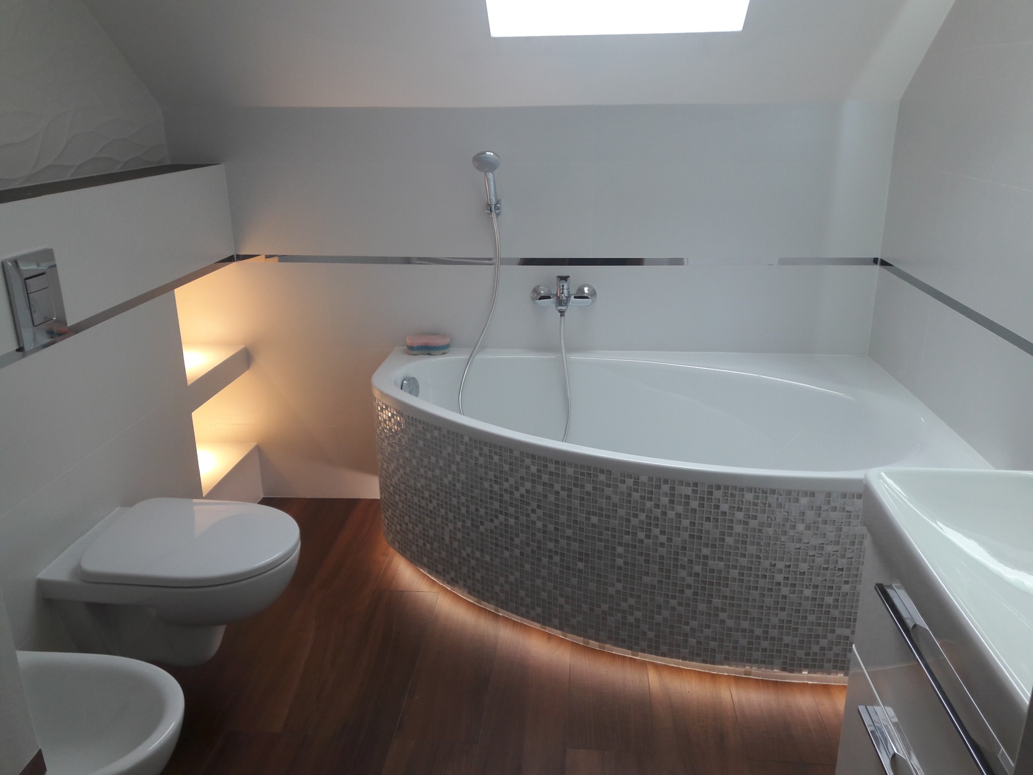 Jasna łazienka na poddaszu z mozaikową obudową wanny podświetloną LED, wiszącą toaletą i bidetem. Uwagę przykuwa skos sufitu i okno dachowe.
