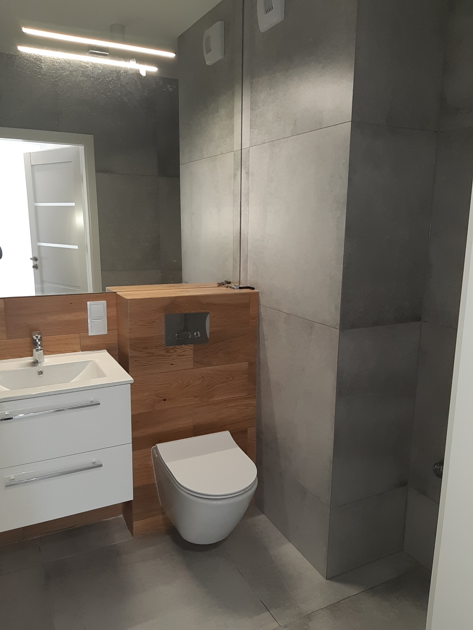 Nowoczesna łazienka z szarymi płytkami, drewnianą zabudową WC i białą szafką z umywalką. Lustro z oświetleniem LED odbija fragment wnętrza.