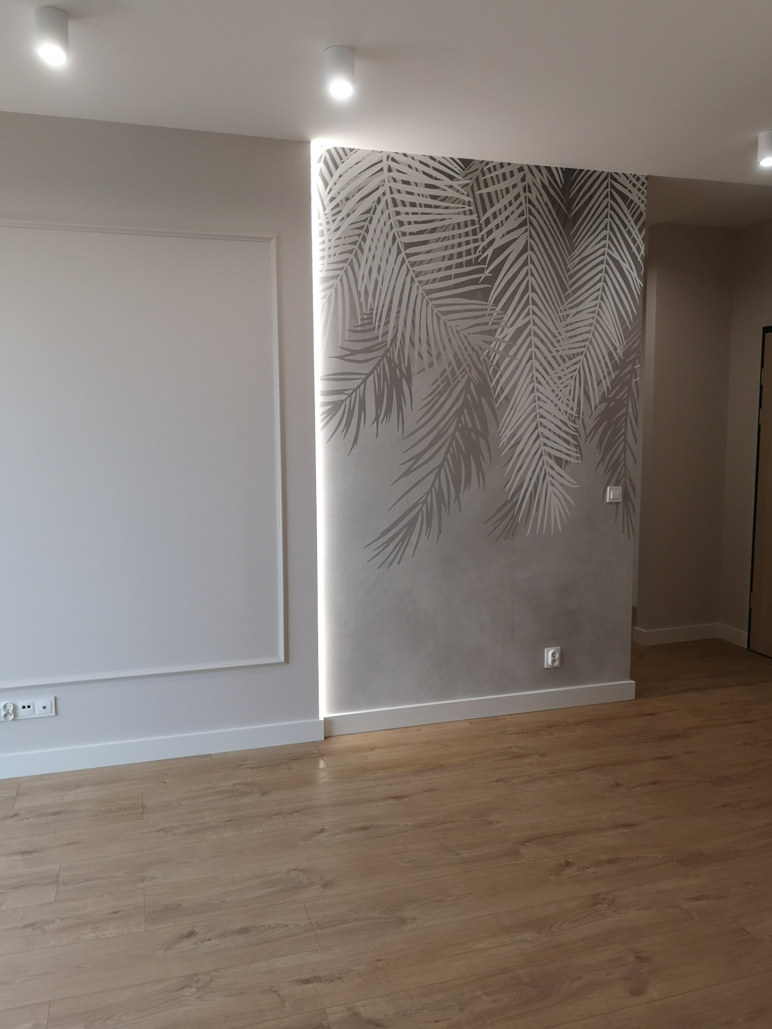 Wykończone wnętrze: gładka, biała ściana z dekoracyjną ramą i fragment ściany z szarą tapetą w motyw liści palmowych, drewniana podłoga, nowoczesne oświetlenie.