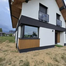 Nowoczesna elewacja domu z białym tynkiem, czarnymi akcentami i drewnianymi panelami pod oknem. Widoczne balustrady balkonowe i kamera monitoringu.