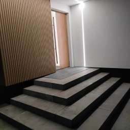 STUDIO P5 Studio Architektury Krajobrazu - Nowoczesne schody wejściowe z kostki brukowej, drewniana okładzina ścienna i subtelne oświetlenie LED tworzą eleganckie wejście do budynku.