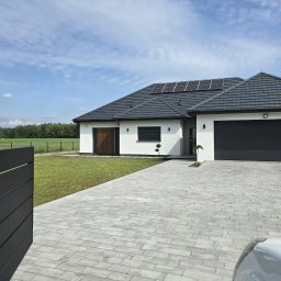 STUDIO P5 Studio Architektury Krajobrazu - Nowoczesny dom jednorodzinny z panelami słonecznymi na dachu, szarą kostką brukową i minimalistycznym ogrodzeniem w tle, widok z poziomu bruku.