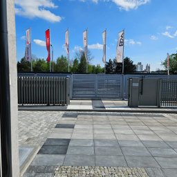 STUDIO P5 Studio Architektury Krajobrazu - Nowoczesne ogrodzenie panelowe w odcieniach szarości, z bramą i furtką, otoczone kostką brukową w różnych wzorach i kolorach. W tle widoczne flagi firmowe i błękitne niebo.