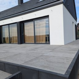 STUDIO P5 Studio Architektury Krajobrazu - Nowoczesny dom z tarasem wyłożonym szarą kostką brukową. Elewacja biało-czarna, duże okna przesuwne. Widoczne detale wykończenia i minimalistyczny styl.