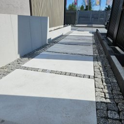 STUDIO P5 Studio Architektury Krajobrazu - Nowoczesna ścieżka z jasnych płyt betonowych, obramowana szarą kostką i drobnym żwirem. W tle budynek z elementami drewnianymi i metalowe ogrodzenie.