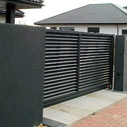 STUDIO P5 Studio Architektury Krajobrazu - Nowoczesna, przesuwna brama lamelowa w kolorze grafitowym, wkomponowana w ciemny, teksturowany mur. Czerwona kostka brukowa i dom z szarą dachówką w tle.