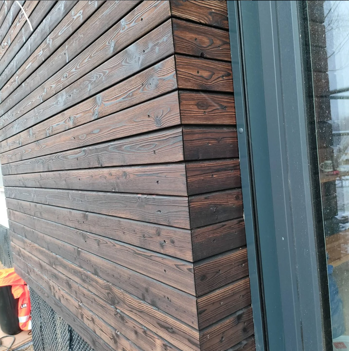 Elewacja z ciemnego drewna, ułożonego poziomo, z widocznymi słojami i fakturą. Szara rama okienna przylega do drewnianej ściany. Widoczne mocowania desek.