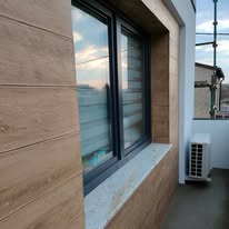 Elewacja budynku w Bełchatowie wykończona panelami imitującymi drewno, z oknem PCV i klimatyzatorem na balkonie. Widoczne rusztowania w tle.