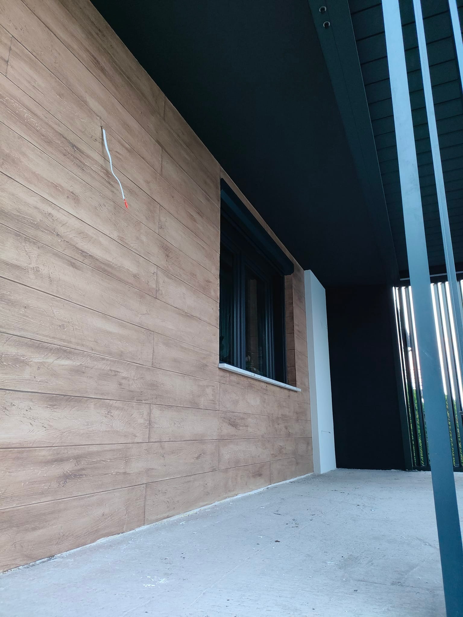 Elewacja budynku z drewnopodobną okładziną, czarną stolarką okienną i fragmentem nowoczesnego ogrodzenia z metalowych profili. Widoczne detale wykończenia.