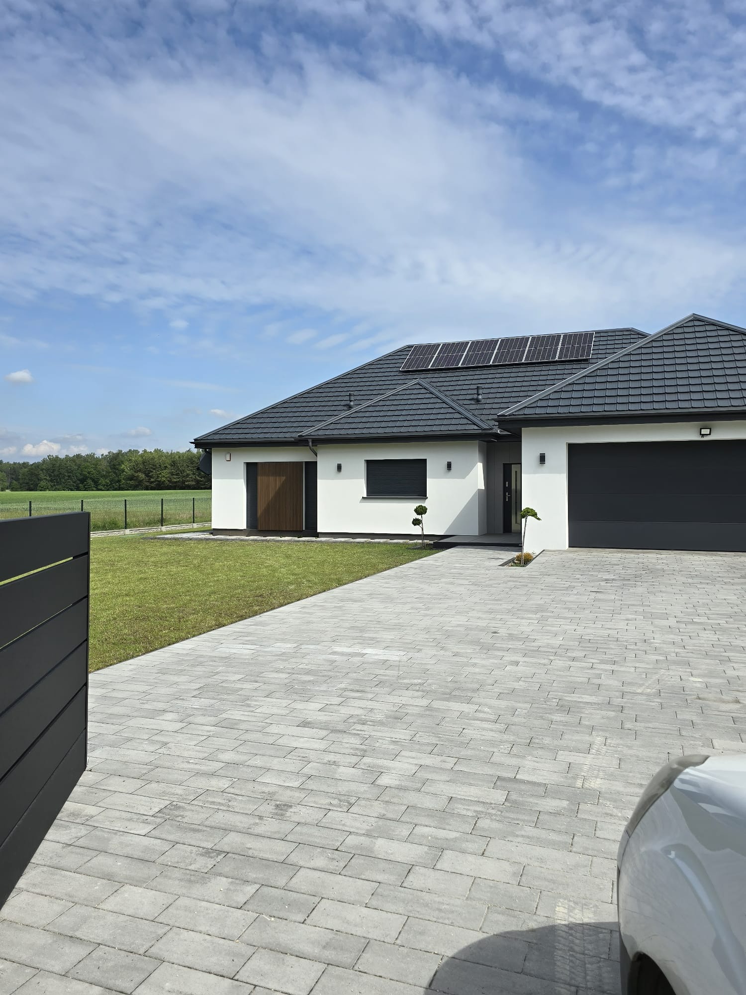 Nowoczesny dom jednorodzinny z panelami słonecznymi na dachu, szarą kostką brukową i minimalistycznym ogrodzeniem w tle, widok z poziomu bruku.