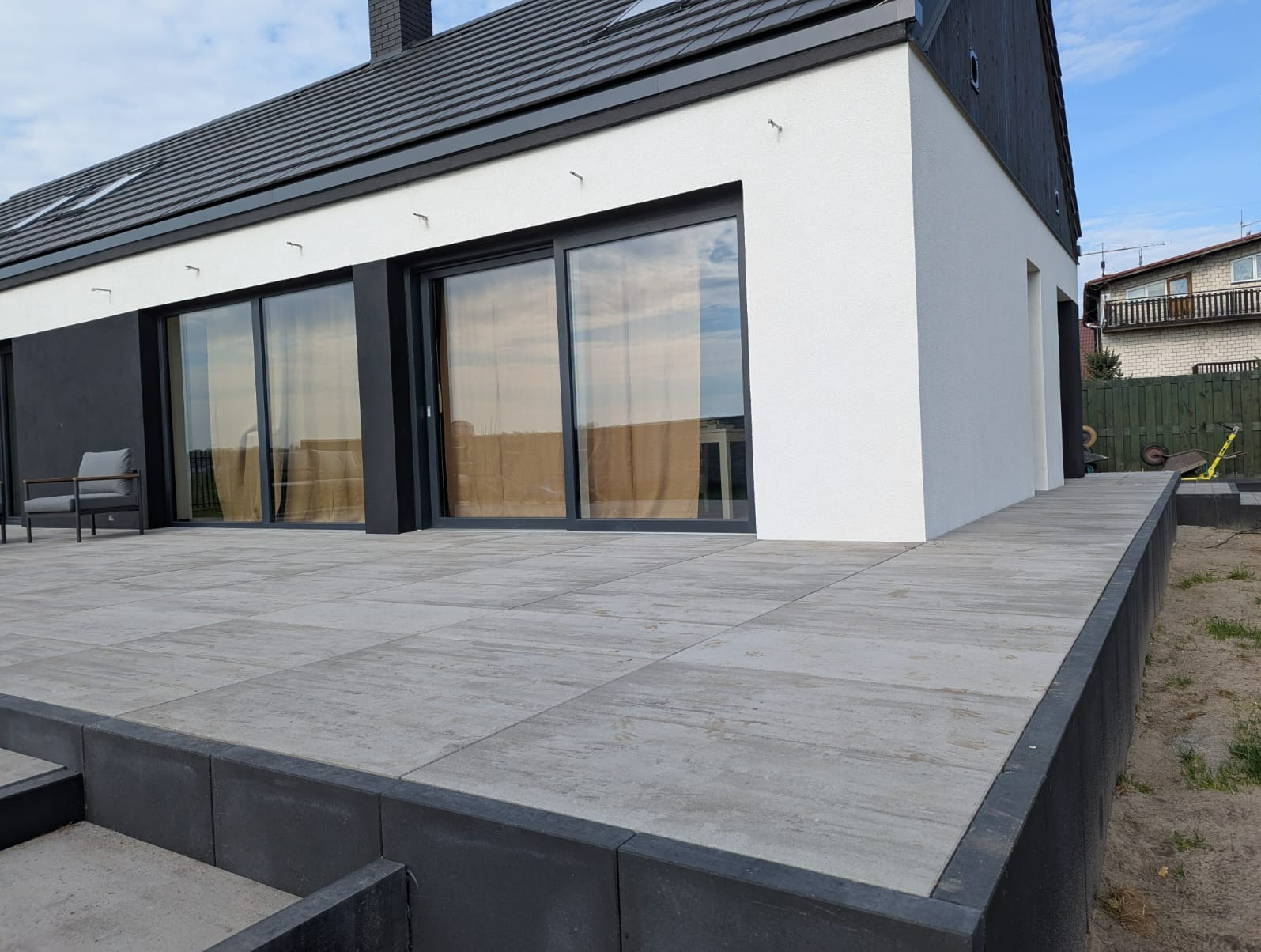 Nowoczesny dom z tarasem wyłożonym szarą kostką brukową. Elewacja biało-czarna, duże okna przesuwne. Widoczne detale wykończenia i minimalistyczny styl.