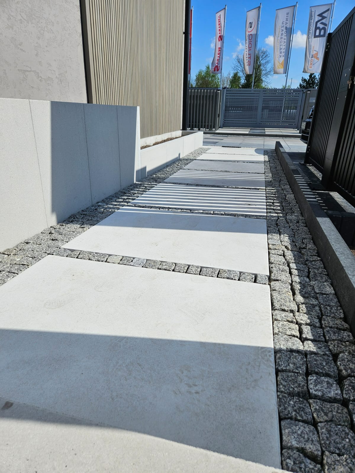 Nowoczesna ścieżka z jasnych płyt betonowych, obramowana szarą kostką i drobnym żwirem. W tle budynek z elementami drewnianymi i metalowe ogrodzenie.