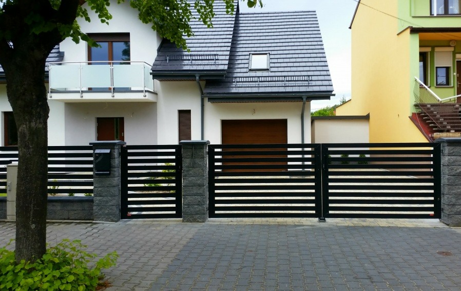 Nowoczesne ogrodzenie panelowe z poziomymi, czarnymi przęsłami i kamiennymi słupkami na tle białego domu z balkonem. Widoczna skrzynka pocztowa i kostka brukowa.