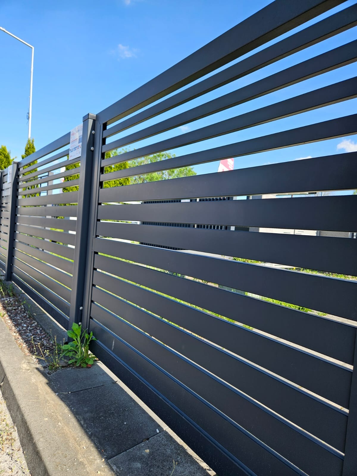 Nowoczesne, poziome ogrodzenie panelowe w antracytowym kolorze, z widocznymi detalami montażu i fragmentem chodnika oraz zieleni w tle.