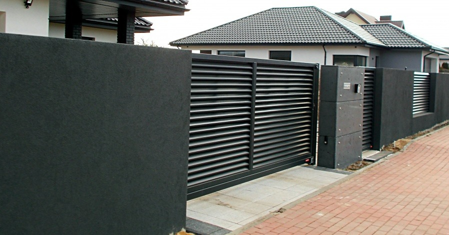Nowoczesna, przesuwna brama lamelowa w kolorze grafitowym, wkomponowana w ciemny, teksturowany mur. Czerwona kostka brukowa i dom z szarą dachówką w tle.