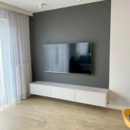 Minimalistyczny salon z wiszącą szafką RTV w bieli, pod telewizorem na szarej ścianie. Jasna podłoga i zasłony w oknie. Nowoczesne oświetlenie sufitowe.