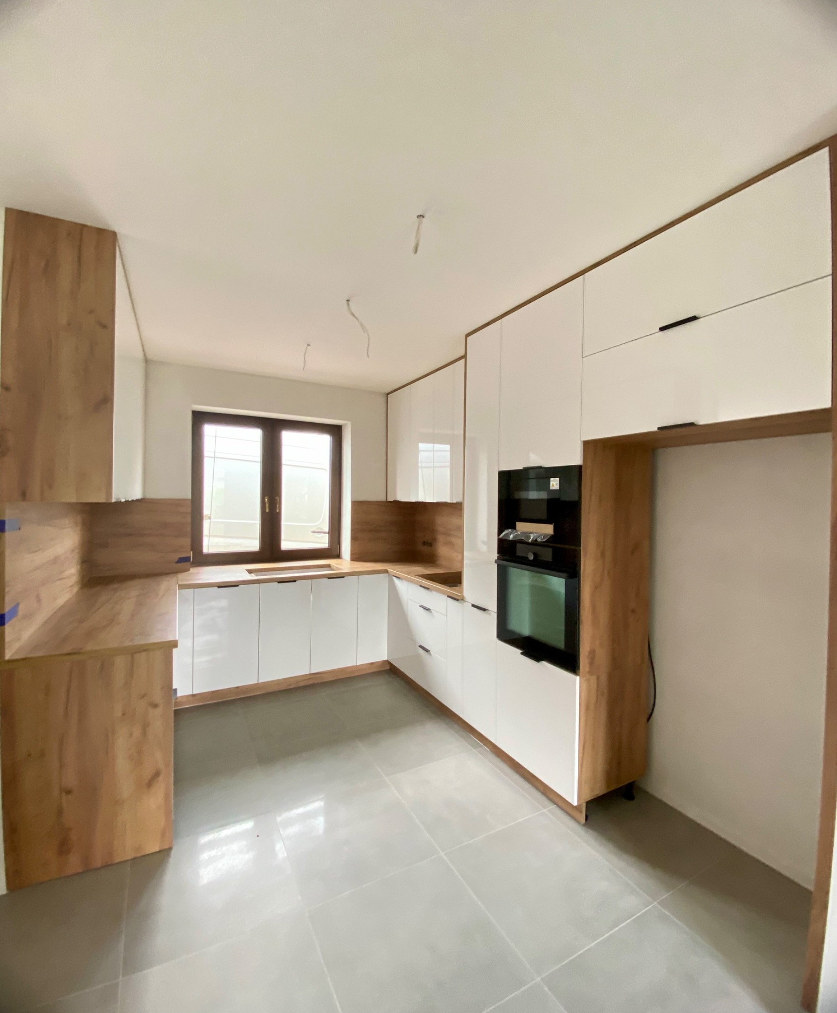 Nowoczesna kuchnia z białymi szafkami i drewnianymi akcentami. Widoczny piekarnik w zabudowie i okno z brązową ramą. Szare płytki na podłodze.