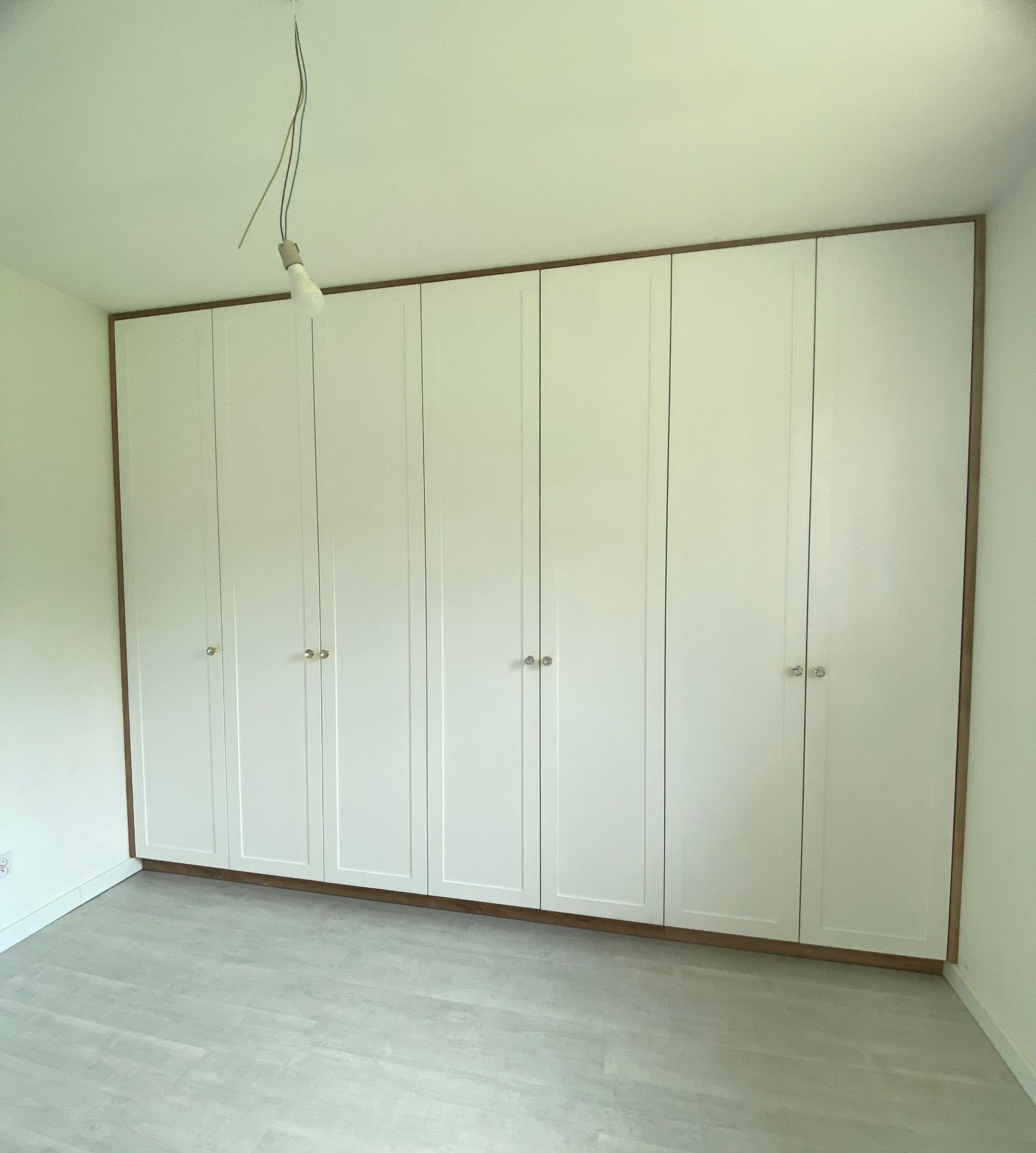 Biała szafa wnękowa z ośmioma drzwiami, obrzeża w kolorze drewna, srebrne gałki, surowa żarówka zwisająca z sufitu. Minimalistyczny design.