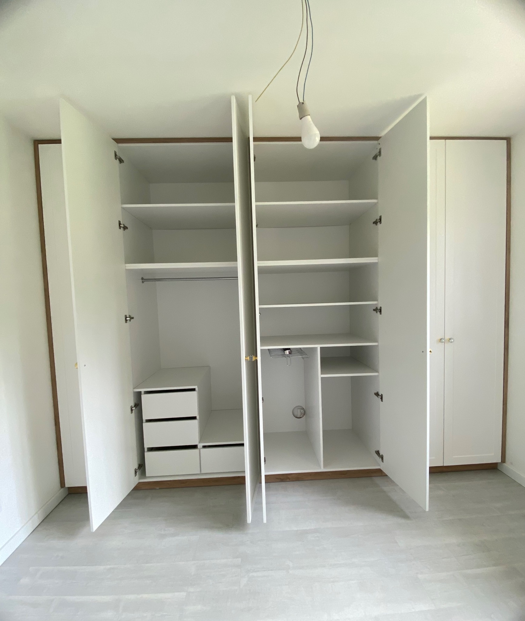 Biała szafa wnękowa z otwartymi drzwiami, widoczne półki, szuflady i drążek na ubrania. U góry żarówka na kablu. Wykończenie w kolorze drewna.