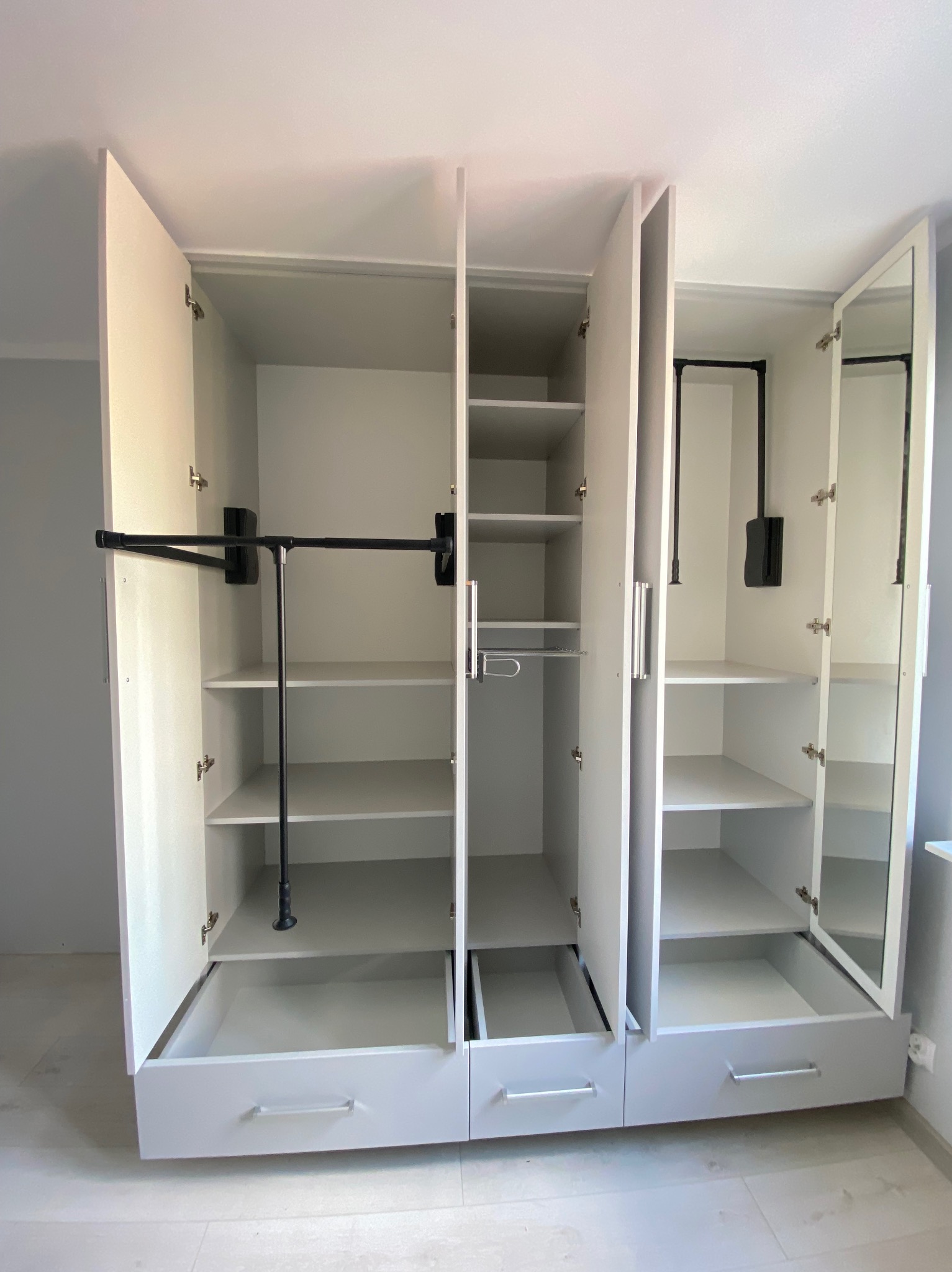 Jasna szafa na wymiar z otwartymi drzwiami, prezentująca półki, szuflady i drążki na ubrania. Widoczne metalowe uchwyty i lustro na jednych z drzwi.