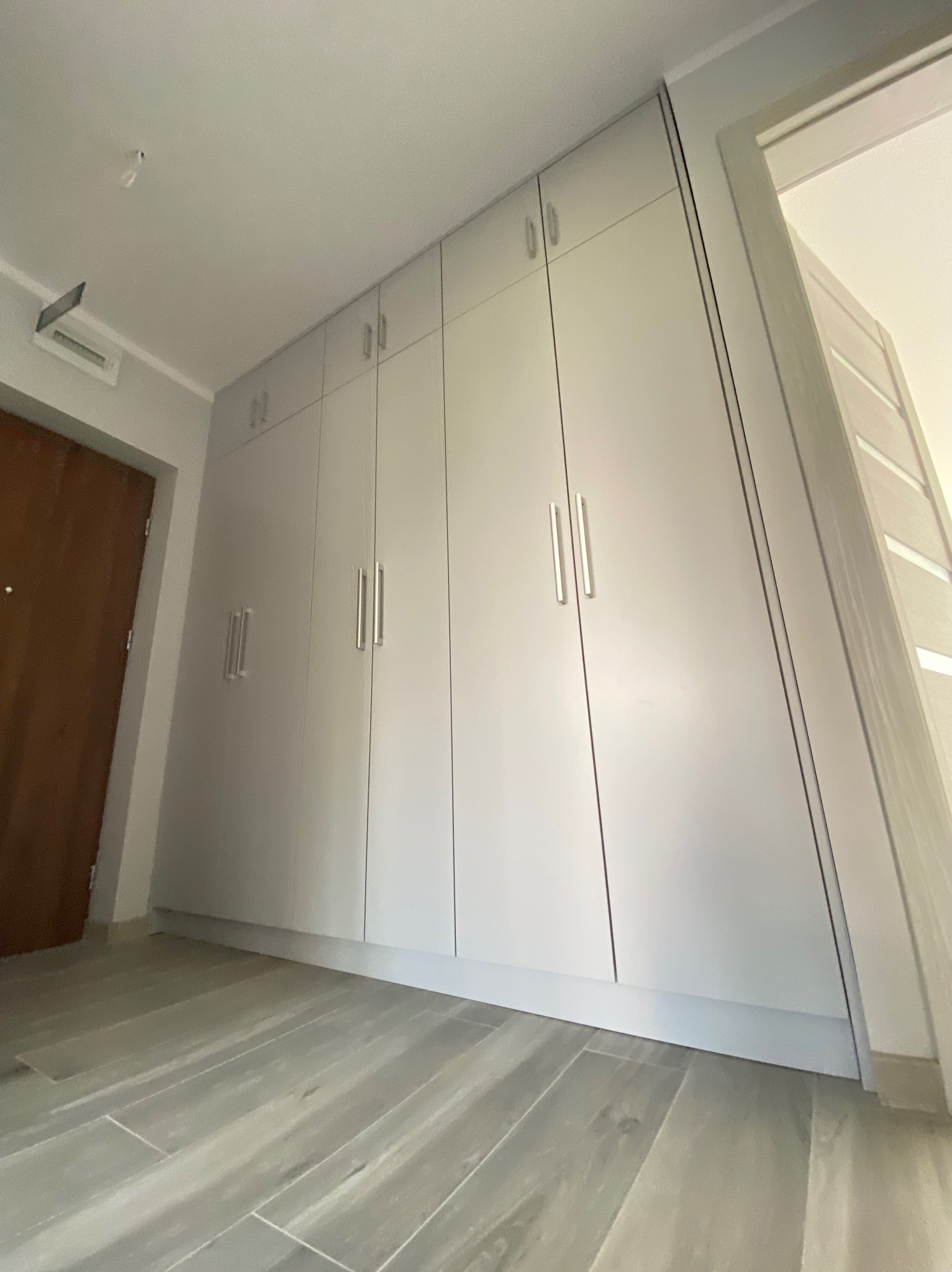 Jasnoszara szafa wnękowa z długimi, srebrnymi uchwytami, wkomponowana w korytarz z szarą podłogą. Widoczne drzwi wejściowe i fragment drugiego pomieszczenia.